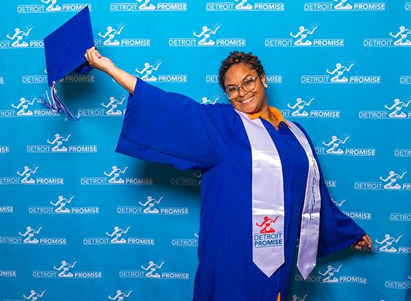 A Detroit Promise graduate in HFC cap and gown holding their cap out in front of a blue Detroit Promise media back drop.