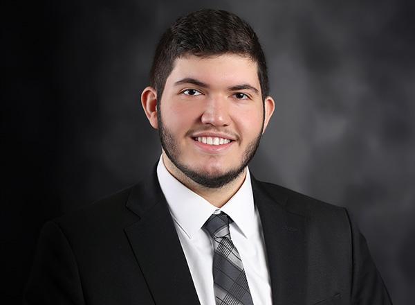 Headshot of Ali Seblini in a black suit with grey plaid tie.