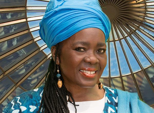 Portrait of Mama Jatu with long hair and earrings, in blue headdress and blue tunic, in front of a large colorful folding fan.
