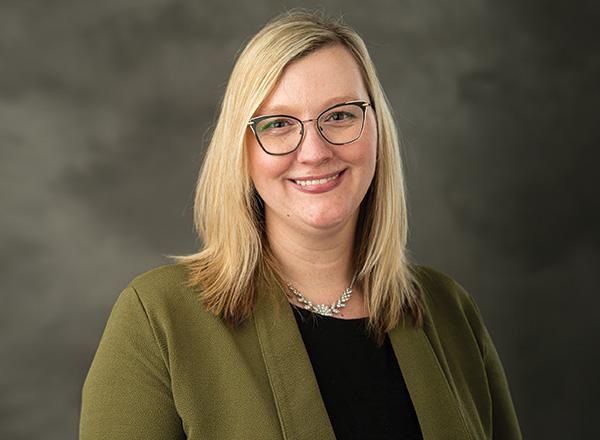 Headshot of Sarah Krizan, who has blonde hair and glasses, wearing an olive green sweater and black shirt.