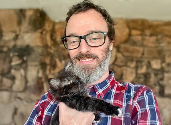 Russell Jones is wearing a plaid shirt and holding a black kitten.