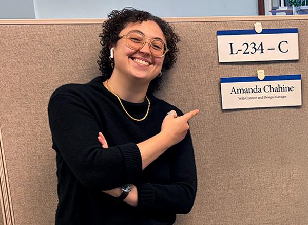 Amanda Chahine poses outside her cubicle, pointing at her name plate.