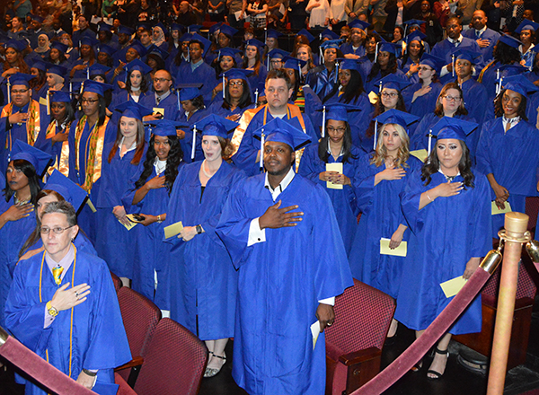 HFC graduates, wearing blue graduation gowns, standing for the National Anthem