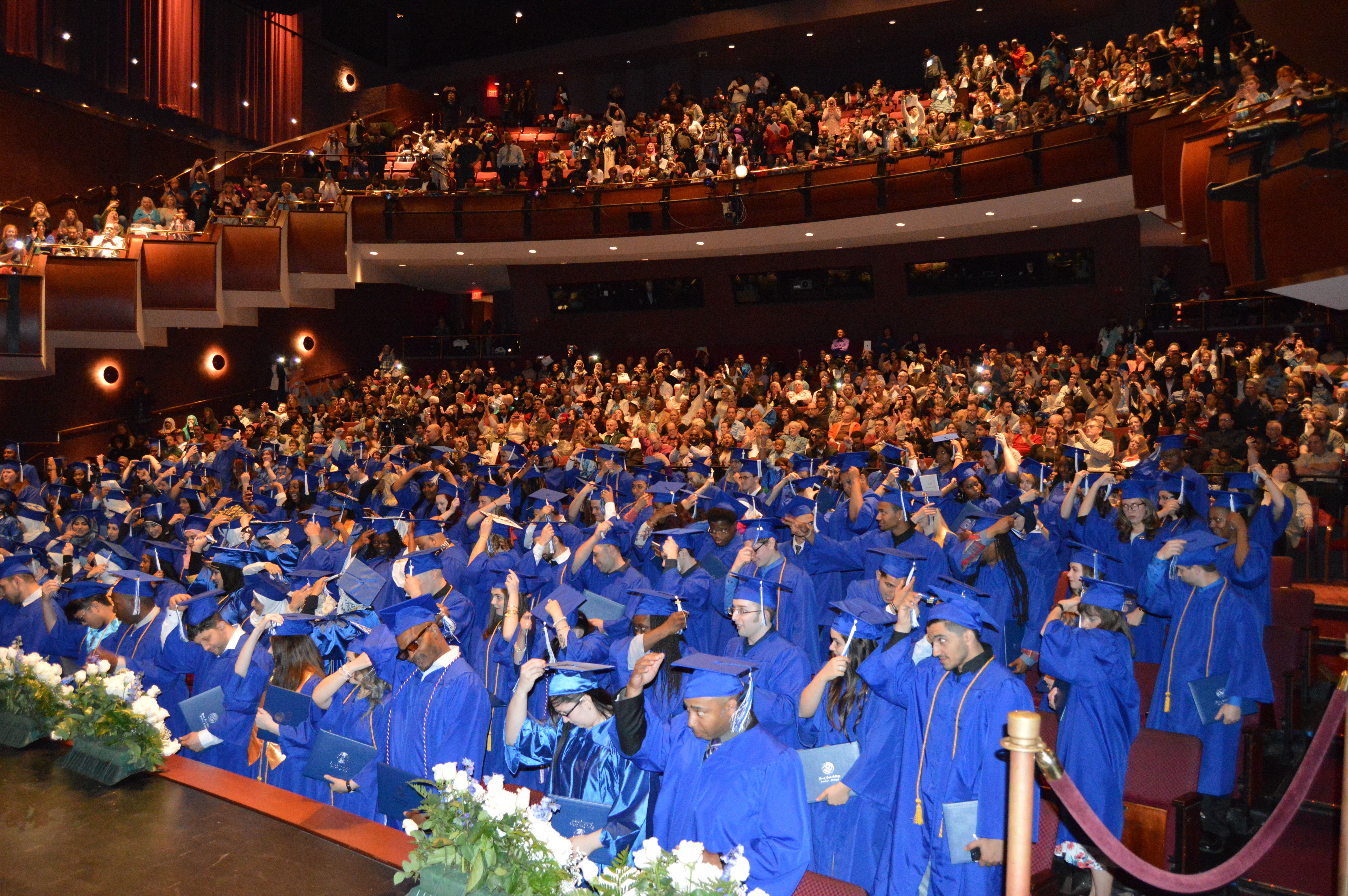 Hundreds of students wearing blue graduation gowns and caps stand in a theatre with their families near the back and on the balcony of the theatre applauding as the students move their tassels.