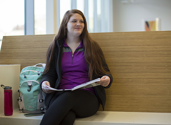 student sits in Science Building lobby, holding notebook and smiling.