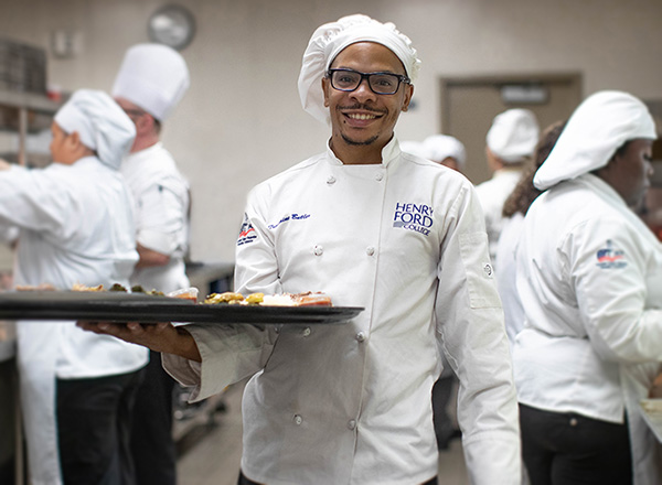 HFC Cuilinary student wearing white chef coat and holding a tray of food. 
